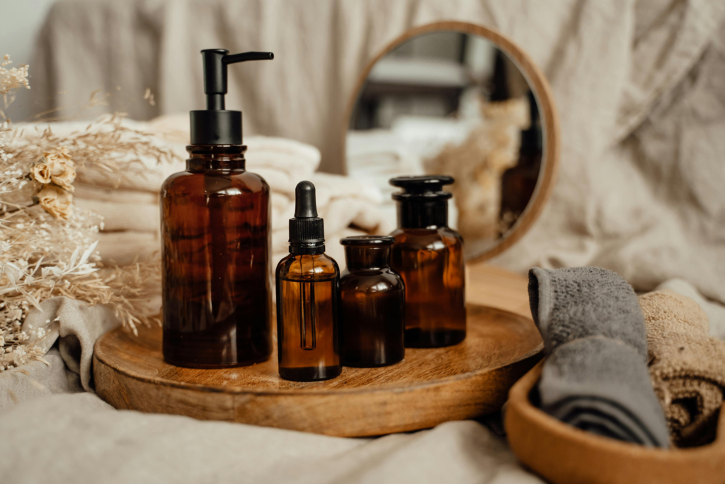 Amber glass skincare and wellness bottles with pump and dropper on a wooden tray beside soft towels and a mirror, creating a calm self-care and relaxation setting.