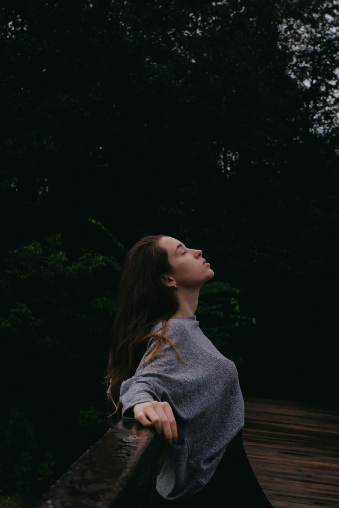 A woman standing with eyes closed head lifted up toward the sky arms stretched out on a wooden reailing in a foresty background