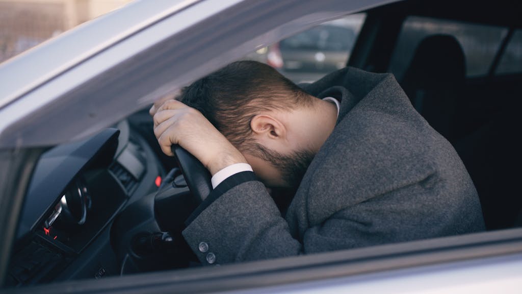 Man feeling overwhelmed and stressed, resting his head on a steering wheel inside a car, stuck in traffic. References survival mode in stressful traffic