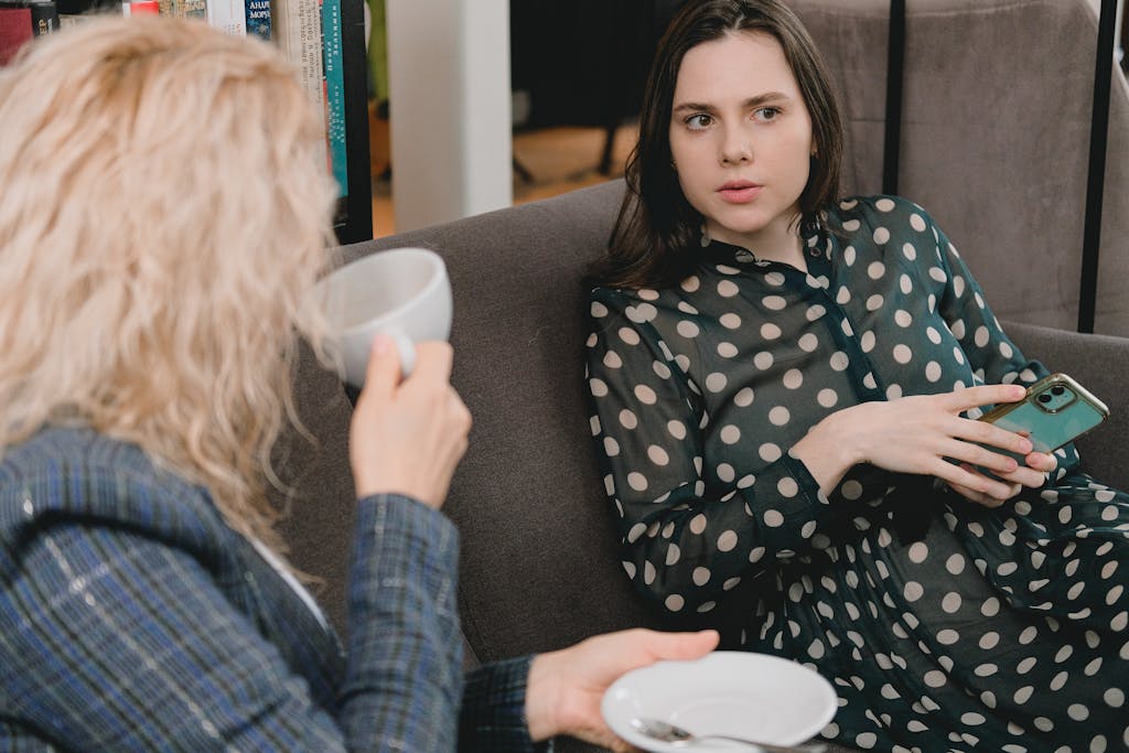 Young female friends in elegant outfits sitting on comfortable sofa and talking during coffee break in cozy cafe