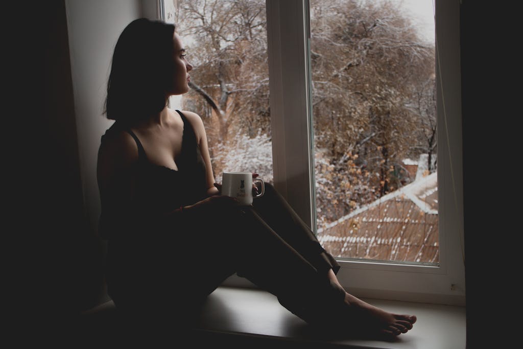 A woman sits by a window, holding a mug, with a wintry scene outside.