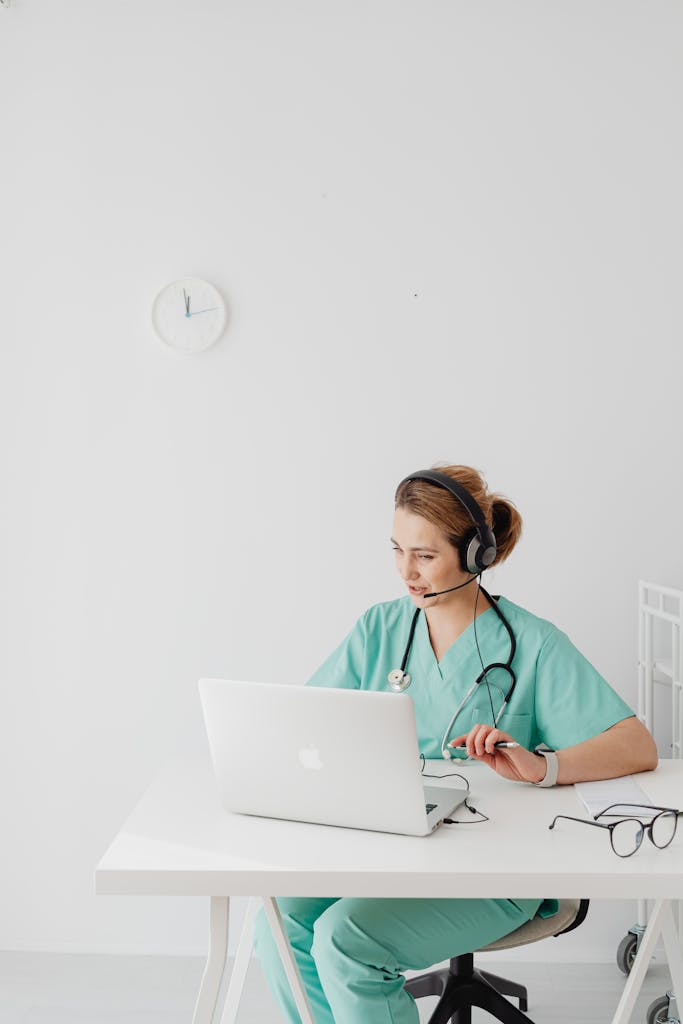 Nurse in teal scrub suit using laptop and headset for virtual consultation in a bright office.