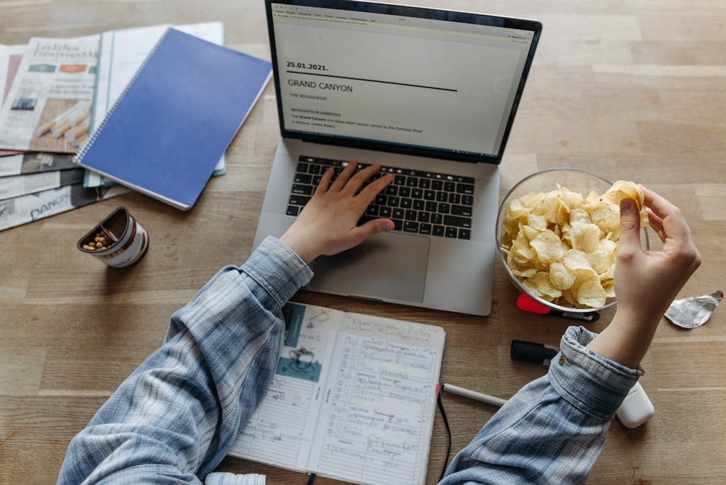 Person multitasking by working on a laptop and eating chips at a desk, blending productivity with leisure.