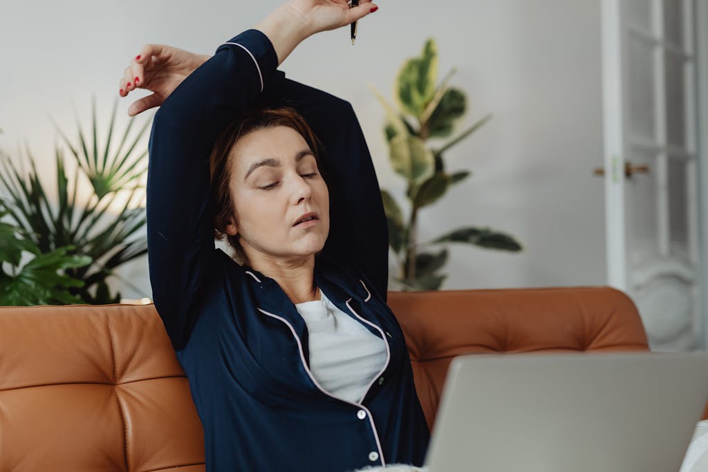 A tired woman in pajamas stretches while working remotely from her living room couch looking exhausted.