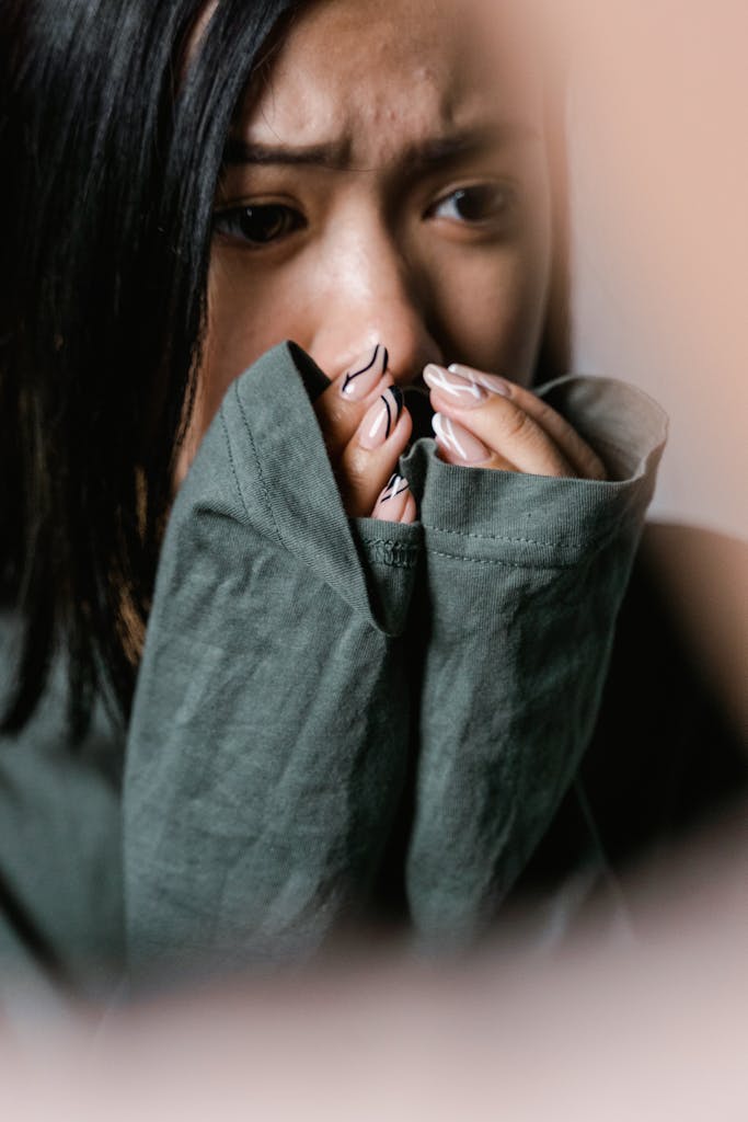 A woman appears physically anxious with symptoms of covering her mouth with a green sweater sleeve in a close-up shot.