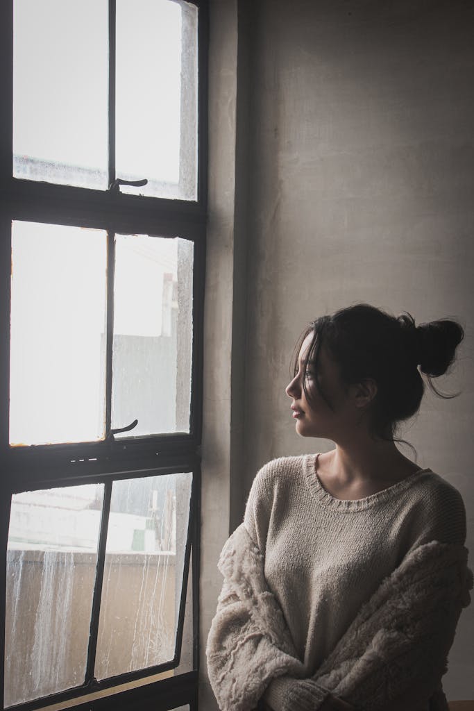 A woman in a cozy sweater looks thoughtfully out a window, creating a serene and contemplative mood.