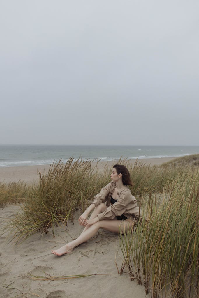 A woman sits on sandy beach among grass, gazing at tranquil ocean horizon under a cloudy sky.