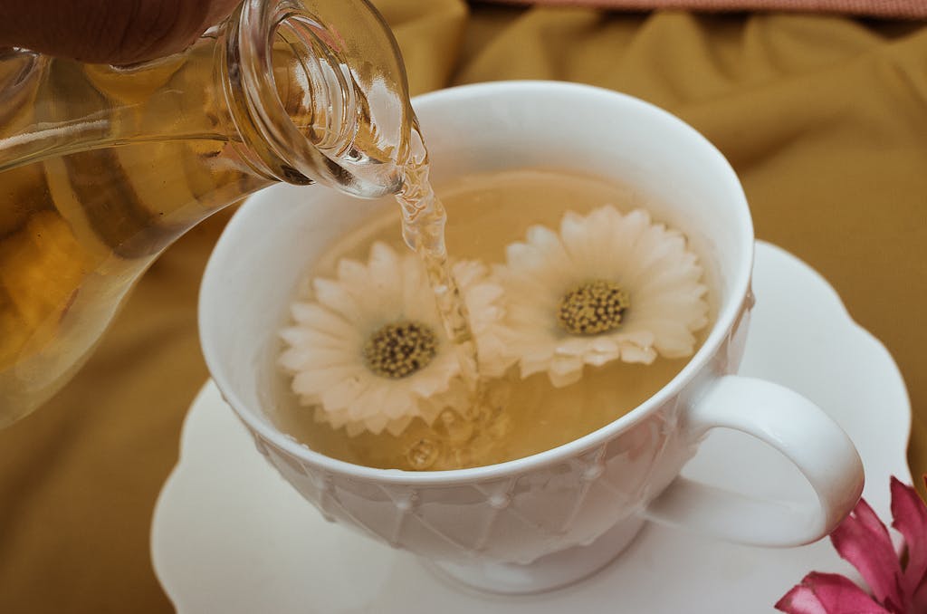 Close-up of herbal tea being poured into a white teacup with floating flowers, creating a serene and relaxing scene.