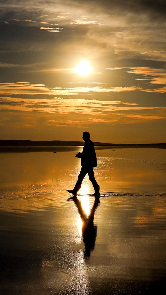 Silhouette of an adult walking on a reflective beach at sunset, creating a dramatic scene.