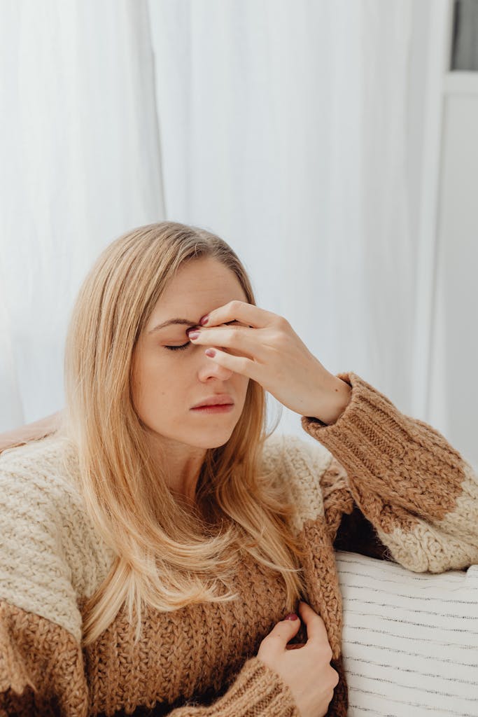 Woman sitting with a headache, wearing a cozy sweater in an indoor setting displaying physical symptoms of pain