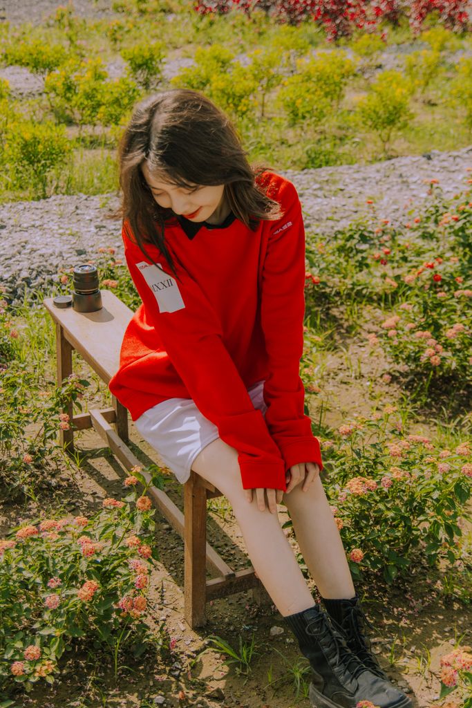 Asian woman in red sweater sits on a bench amidst blooming flowers, enjoying a sunny day resetting her nervous system.