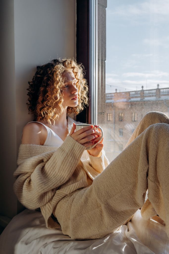 Woman in sweater sits by window, enjoying morning coffee with sunlight calming her nervous system.